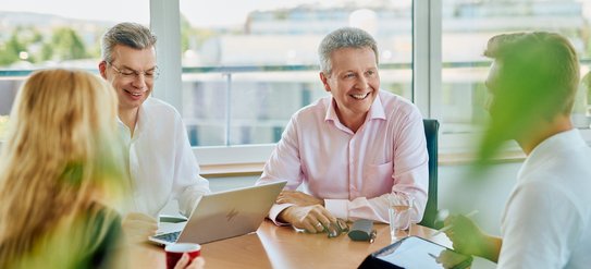 An einem Tisch sitze vier Personen, drei Männer und eine Frau sind in eine Unterhaltung vertieft. Auf dem Tisch steht ein aufgeklappter Laptop.