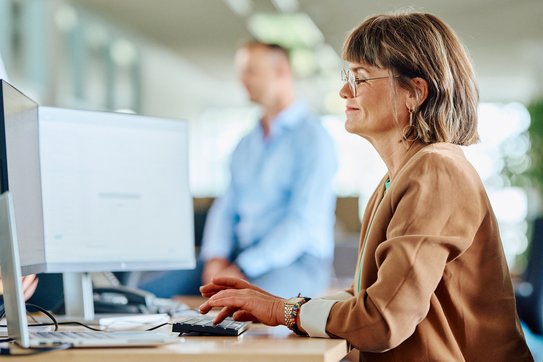 Eine Frau sitzt am Computer, mit zwei Bildschirmen seitlich fotografiert