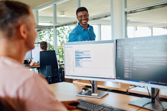 Mitarbeitende unterhalten sich über einen Bildschirm. Auf dem Bildschirm sind Codezeilen zu erkennen.