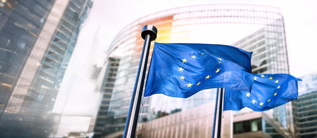 Im Vordergrund sieht man zwei Fahnen mit dem Motiv der EU wehen - Blau mit gelben Sternen. Die Fahnen stehen vor einem Gebäude mit einer großen Glasfassade.