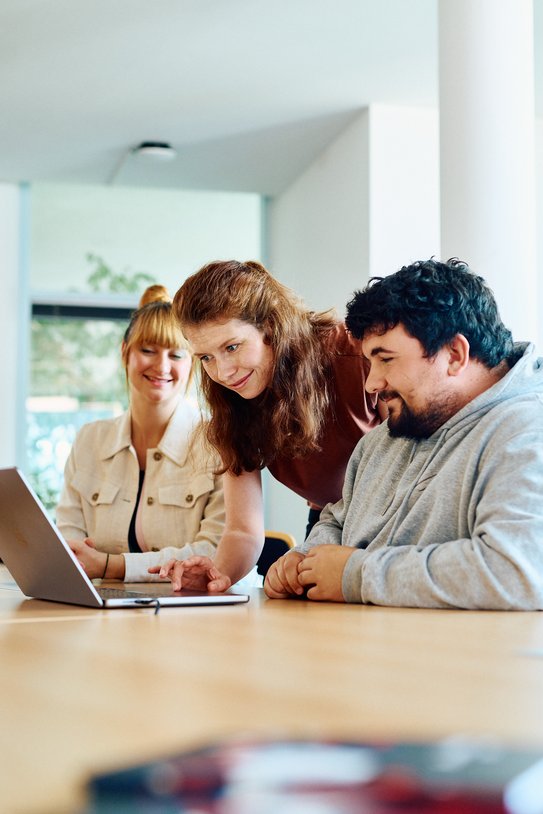 Eine Mitarbeitende zeigt zwei anderen Personen auf dem Laptop etwas, sie steht zwischen den beiden sitzenden Personen. Es wirkt wie eine Schulungssituation.