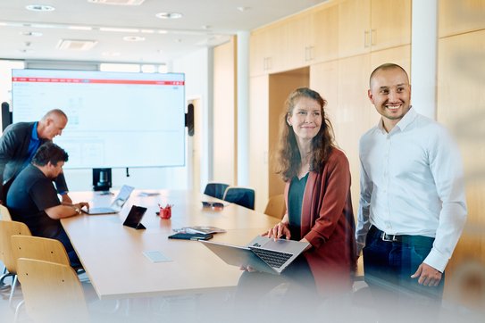 Mehrere Personen befinden sich in einem Besprechungsraum, zwei Personen lehnen dabei vorne im Fokus auf dem Tisch. Davon hält die Frau einen Laptop in ihrer Hand, sie und der Mann neben ihr schauen in Richtung Kamera. Im Hintergrund sieht man zweu Mitarbeitende, die zusammen in einen Laptop schauen.