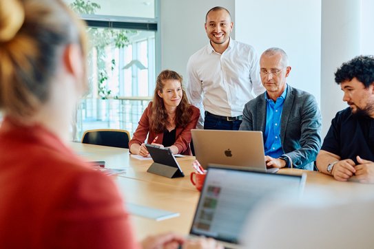 Eine Gruppe Mitarbeotende schauen gemeinsam auf einen Laptop der vor ihnen auf dem Tisch steht