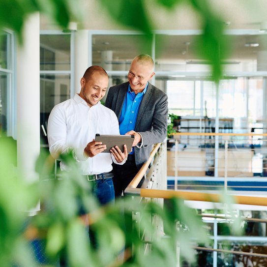 Zwei Mitarbeitende stehen in einem modernen Büro und betrachten gemeinsam ein Tablet.