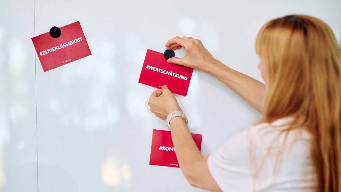 Eine Frau mit langen Haaren hängt eine Postkarte mit dem Wort Wertschätzung an einer Pinnwand auf 