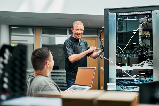 Im Vordergrund sitzt ein Mitarbeiter, welcher nach hinten zu einem weiteren Mitarbeitenden schaut, welcher gerade an einem Serverturm arbeitet