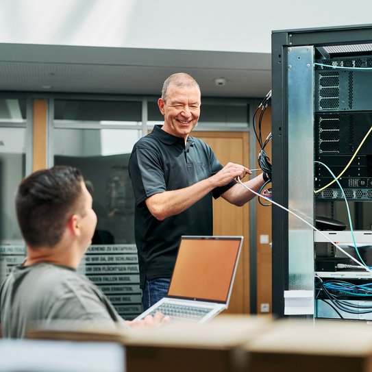 Zwei Mitarbeitende arbeiten gemeinsam an einem Serverturm und ziehen Kabel 