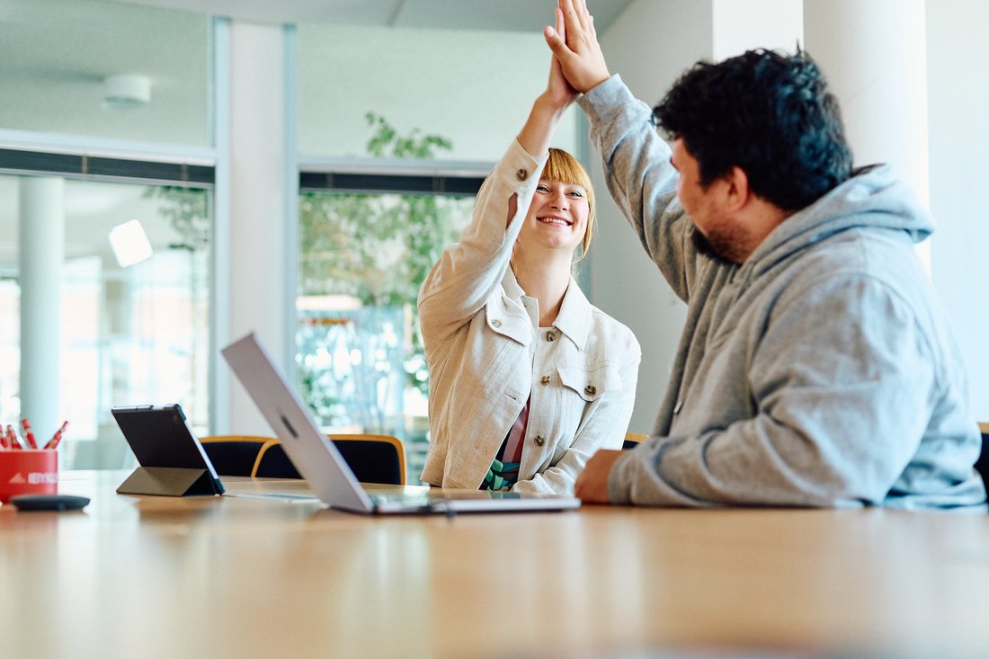 Zwei Mitarbeitende sitzen an einem Tisch und geben sich ein highfive