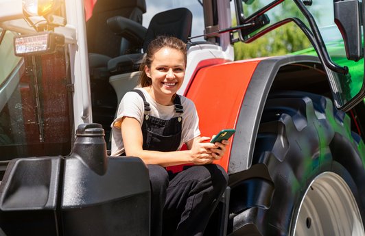 Eine junge Landwirtin sitzt lächelnd auf ihrem Traktor und hält ein Smartphone in den Händen. 