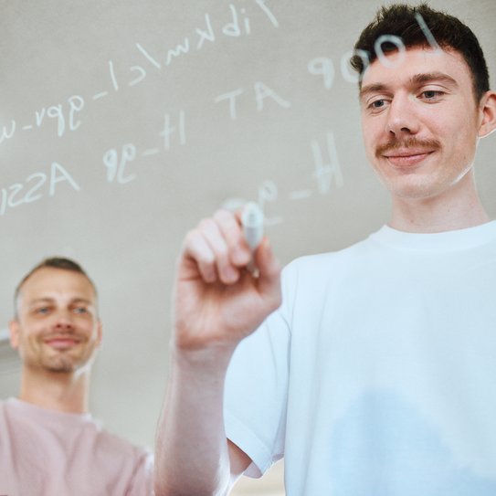 Zwei Mitarbeitende stehen vor einer Glasscheibe auf die sie mit einem weißen Stift Code darauf schreiben