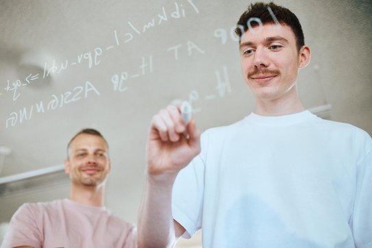 Zwei Mitarbeitende schreiben mit einem Stift Code auf eine Glaswand